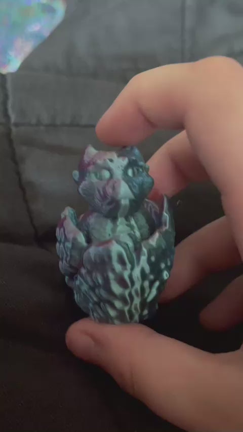 Demonstrating the 2 different noises it can make. There is a hand holding onto the bottom and top and pressing on it down. The item being held is a dragon poking out of an egg. The dragon moves up and down